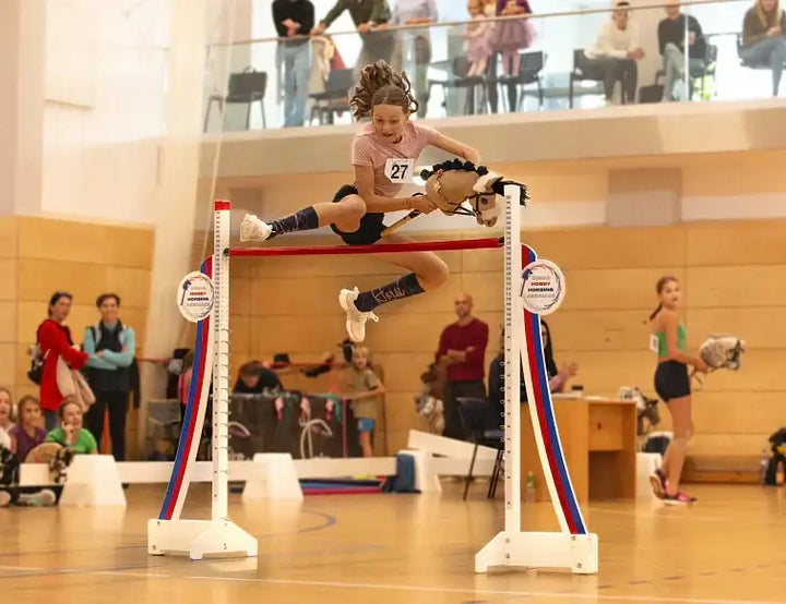 Hobby Horsing competitor jumping over a red pole with a Hobby Horse at an indoor event