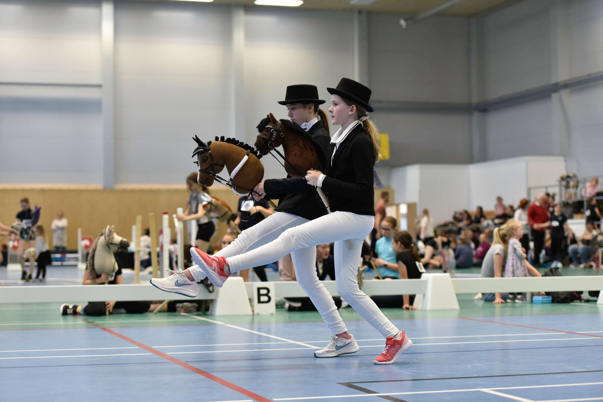 Zwei Kinder in formeller Kleidung gemäß Hobby-Horsing-Kleiderordnung treten mit Hobby Horses in einer Halle an