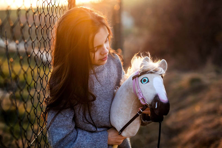 Junges Mädchen hält ein weißes Hobby Horse mit rosa Zaumzeug im warmen Licht des Sonnenuntergangs