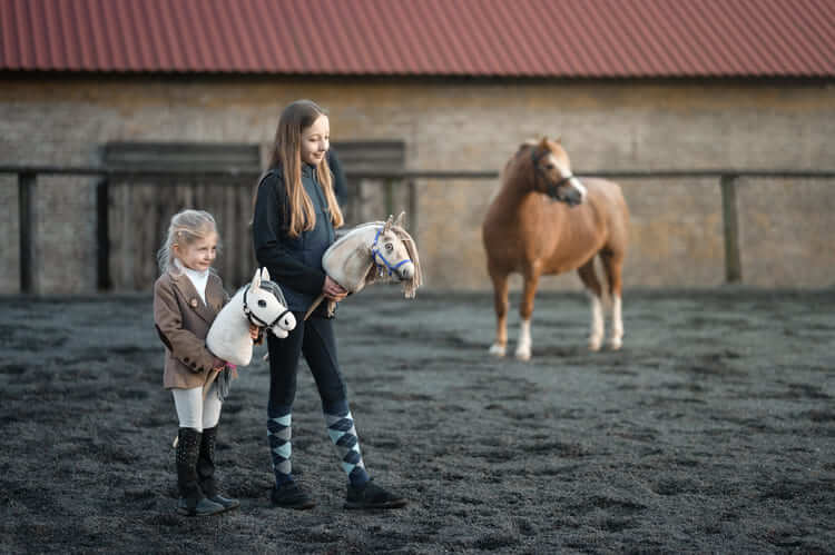Zwei Kinder tragen Hobby Horses neben einem Pony in einer Außenarena