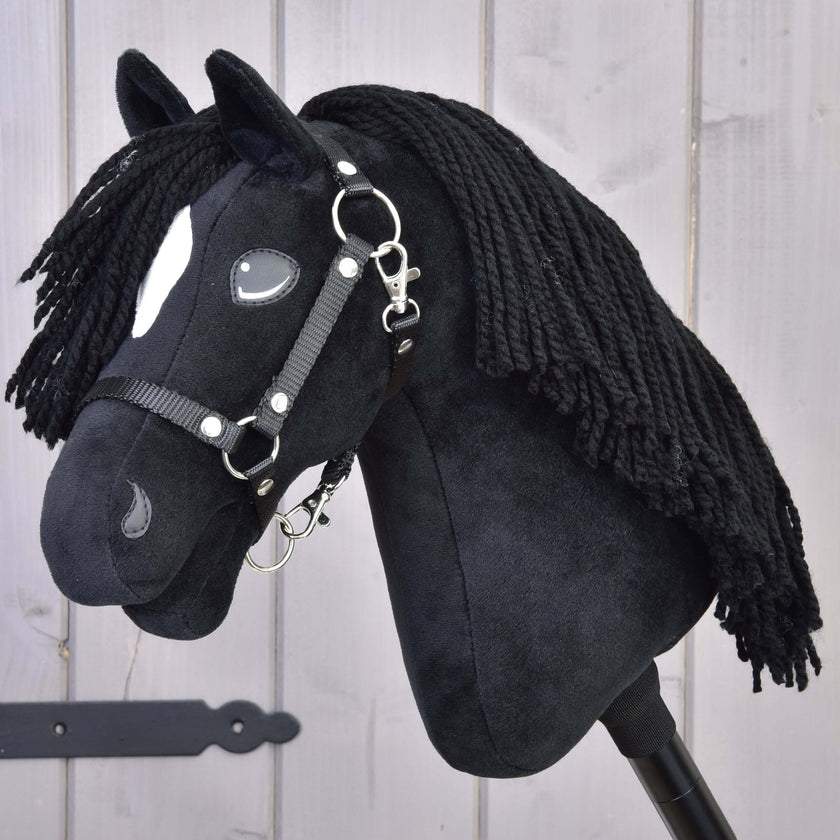 Ein schwarzes Plüsch-Hobby Horse mit langer Garnmähne, weißen Gesichtsabzeichen und schwarzem Zaumzeug auf einem Stock vor hellem Holz-Hintergrund.