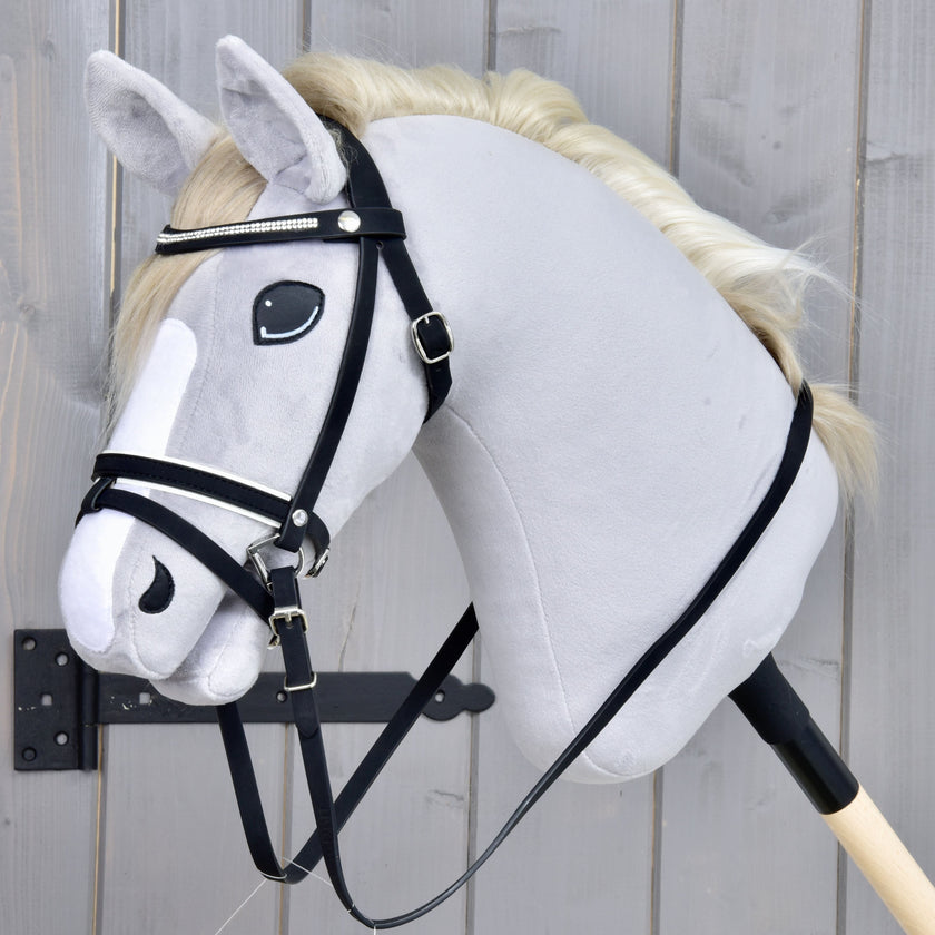 Ein Plüsch-Hobby Horse mit weißer Mähne, schwarzem Zaumzeug und weißer Blesse im Gesicht ist an einem Holzstab befestigt und vor einem grauen Holz-Hintergrund positioniert.