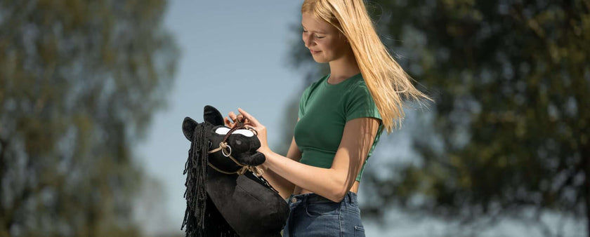 A young woman with long blonde hair, wearing a green t-shirt and jeans, smiles while holding a black hobby horse outdoors on a sunny day, enjoying hobby horsing with trees in the background.