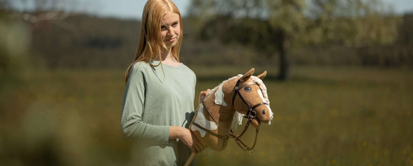 A young woman with long blonde hair stands outdoors in a grassy field, holding a brown and white hobby horse—a nod to the popular hobby horsing trend. Trees and blurred greenery form the backdrop.