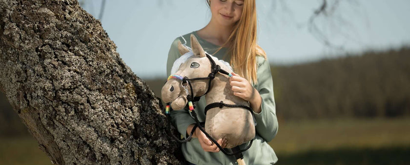 A girl standing outdoors beside a tree holds a handmade hobby horse with a bridle and colorful decorations, smiling gently. The blurred background hints at grassy fields and clear sky, capturing the joy of hobby horsing.