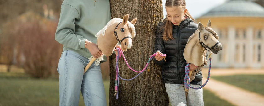 Two children stand outdoors by a tree, each holding a hobby horse toy for a bit of hobby horsing fun. One wears a green sweatshirt and jeans; the other, a black jacket and grey pants. A large building rises in the background.