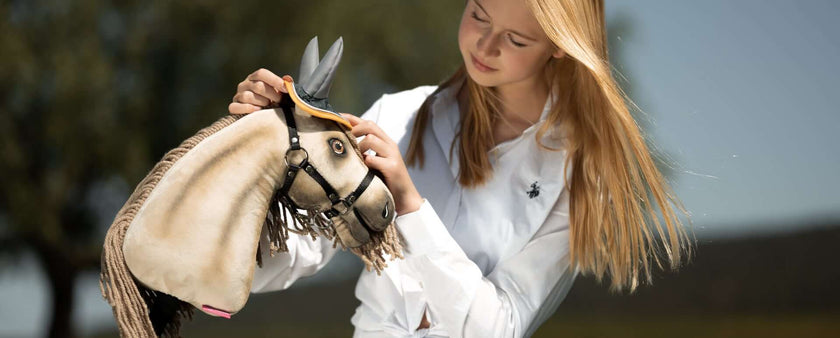 A young girl with blonde hair in a white shirt gently places a ribbon on the head of her realistic Hobby Horse outdoors on a sunny day, capturing the joyful spirit of Hobby Horsing.