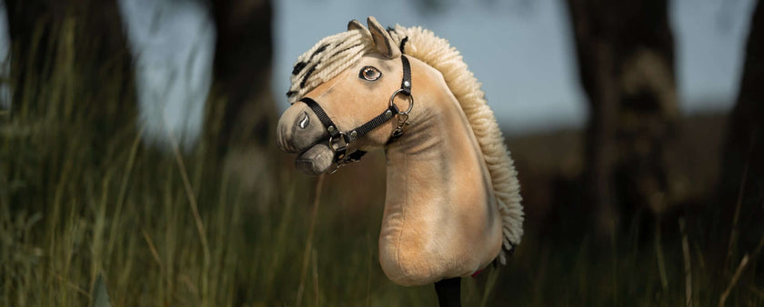 A beige Hobby Horse with a black bridle and white mane stands upright in tall grass, evoking the playful spirit of Hobby Horsing against a backdrop of blurred trees.
