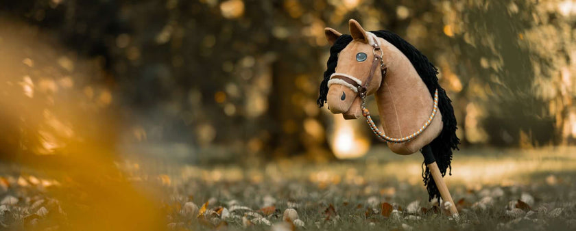 A Hobby Horse with a bridle and black mane stands outdoors on a grassy area, surrounded by fallen leaves—a perfect scene for Hobby Horsing against the blurred natural background.