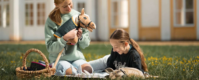 Two girls sit on the grass outside, smiling as they enjoy a sunny day of Hobby Horsing. One holds a hobby horse while the other draws in a notebook. Nearby, a basket and another hobby horse rest on the grass.