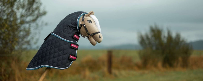 A Hobby Horse with a quilted black blanket and bridle stands outdoors, set against a blurred backdrop of grass, bushes, and overcast sky—perfect for Hobby Horsing adventures.