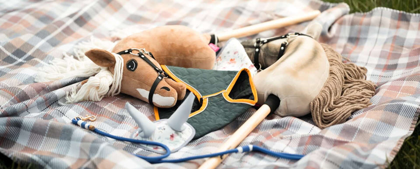Two Hobby Horses and accessories, including a horse mask and a blue lead rope, rest on a plaid blanket spread out on the grass—perfect for an afternoon of Hobby Horsing fun.