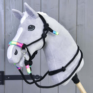 Ein graues Plüsch-Hobby Horse von Hobby Horse LarDen mit weißer Blesse und Mähne, das das Halfter aus dem Seilset Regenbogen trägt, auf einem Holzstab vor einem Hintergrund aus grauen Holzpaneelen.