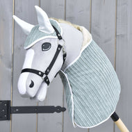 Set Manchester Little Green von Hobby Horse LarDen zeigt ein grauweißes Hobby Horse mit weichem Stoffkopf, schwarzem Zaumzeug, Fliegenhaube und hell blaugrünem Halsteil an einem Holzstab, vor einem Holzzaun.