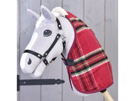 Ein graues Hobby Horse Spielzeug von Hobby Horse LarDen mit Stalldecke Christmas in rotem Karomuster und schwarzem Zaumzeug ist auf einem Holzstab vor einer hellen Holzpaneel-Kulisse.