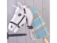 Hobby Horse en peluche de Hobby Horse LarDen, avec visage gris et blanc, bride noire et couverture d'écurie écossaise à carreaux bleue, monté sur un bâton devant un mur en bois clair.