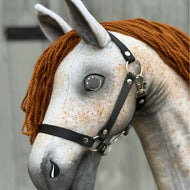 Hobby Horse Pompo mit schwarzem Halfter von Hobby Horse LarDen, Nahaufnahme mit handgemachtem grauem Körper, Knopfaugen, brauner Garnmähne und schwarzem Halfter, die den verspielten Look eines Hannoveraners vor verschwommenem Holzhintergrund zeigt.