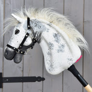 Hobby Horse Aspen mit schwarzem Halfter, Größe S von Hobby Horse LarDen mit weißem Plüschkopf mit schwarzen Flecken, flauschiger Mähne und schwarzem Halfter, auf einem Holzstab montiert vor einer grauen Holzwand