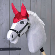 Das graue Hobby Horse von Hobby Horse LarDen mit weißer Mähne, schwarzem Zaumzeug und Ohrenkappe gehäkelt rot zum Fliegenschutz steht vor einem hellen Holz-Hintergrund.