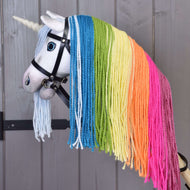 The Hobby Horse LarDen Hobby Horse Unicorn Rainbow with Bridle and Halter features a white horn, blue eyes, a bridle, and a vibrant rainbow mane with blue, green, yellow, orange, and pink yarn strands against a wooden background.
