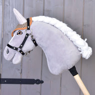 Hobby Horse LarDen en peluche avec tête grise, bride noire, bonnet d’oreilles anglais marron à carreaux pour la protection contre les insectes, crinière blanche, monté sur un bâton en bois devant un fond en panneaux de bois.