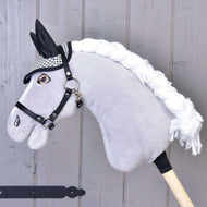 The Hobby Horse LarDen "Ear Bonnet English Black Plaid" is a plush hobby horse with a gray face, white yarn mane, black harness, and plaid ear bonnet for insect protection, mounted on a wooden stick against a gray wall.