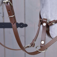 Nahaufnahme des Hobby Horse LarDen Hellbraunes Kunstlederset - Zaumzeug, Zügel und Martingale mit Metallringen und Schnallen auf hellgrauem Stoff vor einem Holzhintergrund mit schwarzem Metallscharnier.