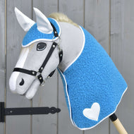 Hobby Horse LarDen „Set Mystery“ mit einem grauen Pferd mit blauer strukturierter deckenartiger Hülle, weißen Ohren, weißem Herz an der Seite, schwarzem Zaumzeug, dichter weißer Mähne und auf einem Stab vor Holzhintergrund.
