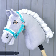 Ein grau-weißes Hobby Horse mit geflochtener weißer Mähne trägt das Hobby Horse LarDen „Halfter türkis mit Fell“, das perfekte Accessoire, vor hellgrauem Holz-Hintergrund.