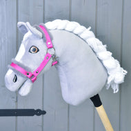 Hobby Horse LarDen „Halfter pink mit Fell“ auf einem grauen Hobby Horse mit weißer Garnmähne, weißen Gesichtsabzeichen und Holzstab vor grauem Holzhintergrund.