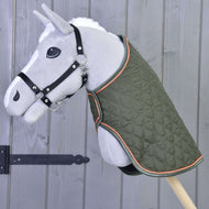 Ein graues Plüsch-Hobby Horse auf einem Stab mit der Hobby Horse LarDen Decke Sport in Grün mit orangefarbener Einfassung und einem schwarzen Halfter für Hobby Horse steht vor einer Holzwand mit schwarzem Eisenscharnier.