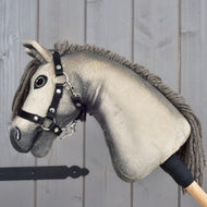 Le Hobby Horse Dusty avec licol noir de LarDen (taille XS) présente une tête hanovrienne en peluche avec des yeux boutons et un licol noir, montée sur un bâton en bois devant un fond de panneaux en bois clair