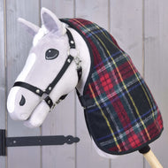 Ein Plüsch-Hobby Horse mit weißem Gesicht und schwarzem Zaumzeug trägt die Hobby Horse LarDen Stalldecke Edinburgh in rot-grünem Karomuster und ist vor einer grauen Holzwand mit sichtbarem schwarzem Metallscharnier positioniert.