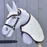 Das Set Leonardo von Hobby Horse LarDen zeigt ein graues Plüsch-Hobby Horse mit weißer gesteppter Decke, passender Fliegenhaube, schwarzem Zaumzeug und Zügeln auf einem Holzstab vor einer grauen Holzwand.