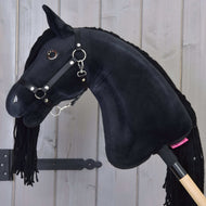 Hobby Horse Maximus mit Schwarzem Halfter von Hobby Horse LarDen mit glänzenden Knopfaugen und Garnmähne, auf einem Holzstab, vor einer Wand aus hellen Holzplanken.