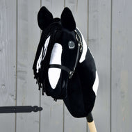 Hobby Horse Merlin mit Schwarzem Halfter von Hobby Horse LarDen ist ein schwarz-weiß geflecktes Hobby Horse nach dem Vorbild eines Barock Pinto Hengsts, mit Garnmähne und Zaumzeug, auf einem Holzstab vor einem grauen Zaun.