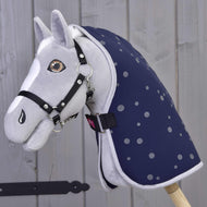 Hobby Horse en peluche gris de Hobby Horse LarDen avec bride noire et Couverture d'écurie Ciel de Nuit, monté sur un bâton en bois devant un mur en bois clair.