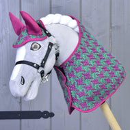 Un Hobby Horse gris de Hobby Horse LarDen se tient devant un mur en bois gris clair, portant l’Ensemble Coco : un ensemble couverture à motif pied-de-poule rose et vert avec bonnet d’oreilles rose assorti et bride noire.