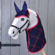Das Hobby Horse LarDen „Set Tartan“ zeigt ein Plüsch-Hobby Horse mit grauem Gesicht, schwarzem Zaumzeug, blauen Ohren und einer dunkelblauen karierten Decke mit rotem Rand, vor einer grauen Holzwand.