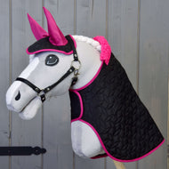 Das Hobby Horse LarDen Set Thermo schwarz mit Pelz zeigt ein plüschiges Hobby Horse mit weißem Gesicht, pinken Ohren, schwarzem Zaumzeug und geflochtener weißer Mähne vor Holzhintergrund. Es hat eine schwarze gesteppte Decke mit leuchtend pinkem Besatz.