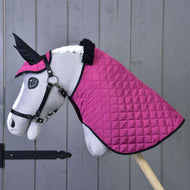 Ein plüschiges Hobby Horse von Hobby Horse LarDen steht auf einem Holzstab, mit weißem Gesicht, schwarzem Zaumzeug und dem Set Thermo Pink mit Decke und Ohrenkappe vor einer grauen Wand.