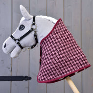 Ensemble festif Bordeaux de Hobby Horse LarDen, Hobby Horse gris en peluche avec bride noire et couverture pied-de-poule bordeaux et rose, monté sur un bâton en bois comme décoration charmante devant un fond en bois gris.