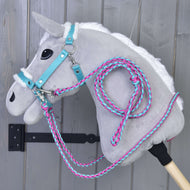 Set Perfect Combination Turquoise von Hobby Horse LarDen, ein plüschiges graues Hobby Horse mit weißer Mähne, türkisfarbenem Zaumzeug für Hobby Horse, mit Fell gepolstertem Halfter für Hobby Horse und pink-blauen Zügeln aus Seil, hängt vor einer grauen Holzwand.