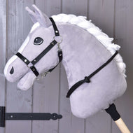 Halsring schwarz von Hobby Horse LarDen an einem grauen Plüsch-Hobby Horse mit weißer Mähne auf einem Holzstab, mit schwarzem Zaumzeug für Hobby Horse, Zügeln und handgefertigtem Halsring, vor einer grauen Holzwand.