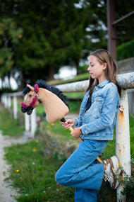 Mädchen im Jeanslook lehnt an einem weißen Koppelzaun und hält das Hobby Horse Cookie mit schwarzem Halfter von Hobby Horse LarDen – sandfarbener Plüschkopf, schwarze Garnmähne – hier mit dem fellgefütterten Teddy Maroon Halfter
