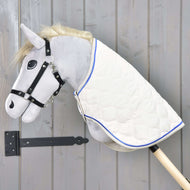 Das Hobby Horse LarDen mit Decke Cream Pearl zeigt einen plüschigen grauen Kopf, schwarzes Zaumzeug und eine cream pearl Decke mit blauem Rand auf einem Holzstock, vor einer grauen Holzwand.