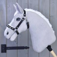 Ein graues Hobby Horse mit weißer Mähne und schwarzem Zaumzeug ist auf einem Holzstab montiert und trägt das Halfter mit Fell- Kunstleder Espresso von Hobby Horse LarDen, vor einer hellgrauen Holzwand mit einem links sichtbaren schwarzen Scharnier.