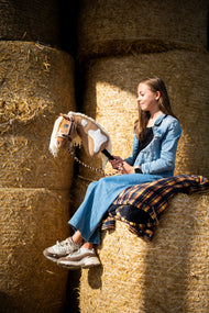 Mädchen in Jeansjacke sitzt hoch auf runden Strohballen und hält das Hobby Horse Fury mit schwarzem Halfter von Hobby Horse LarDen – Palomino-Plüschkopf mit weißen Abzeichen, cremefarbene Garnmähne – hier mit dem fellgefütterten LarDen Brown Halfter und dem Bloom Brown Führstrick