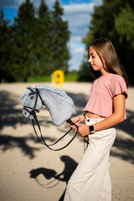 Mädchen im rosa Oberteil führt das Hobby Horse Grace mit schwarzem Halfter von Hobby Horse LarDen über einen Sandplatz – grauer Plüschkopf, hellgraue Garnmähne – hier mit dem vollständigen De Luxe Zaumzeug mit zwei Stirnbändern
