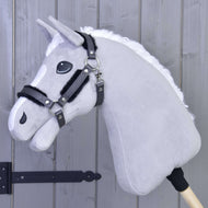 Halfter mit Fell- Kunstleder Graphite Full von Hobby Horse LarDen ist auf einem Holzstab montiert und lehnt an einer hellgrauen Holzwand. Zu sehen sind ein grauer Plüschkopf mit weißer Mähne, schwarzes Zaumzeug und ein Halfter aus Kunstleder.